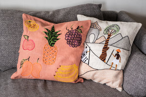Two embroidered pillows on a gray couch, one with fruit patterns and the other with a landscape design.